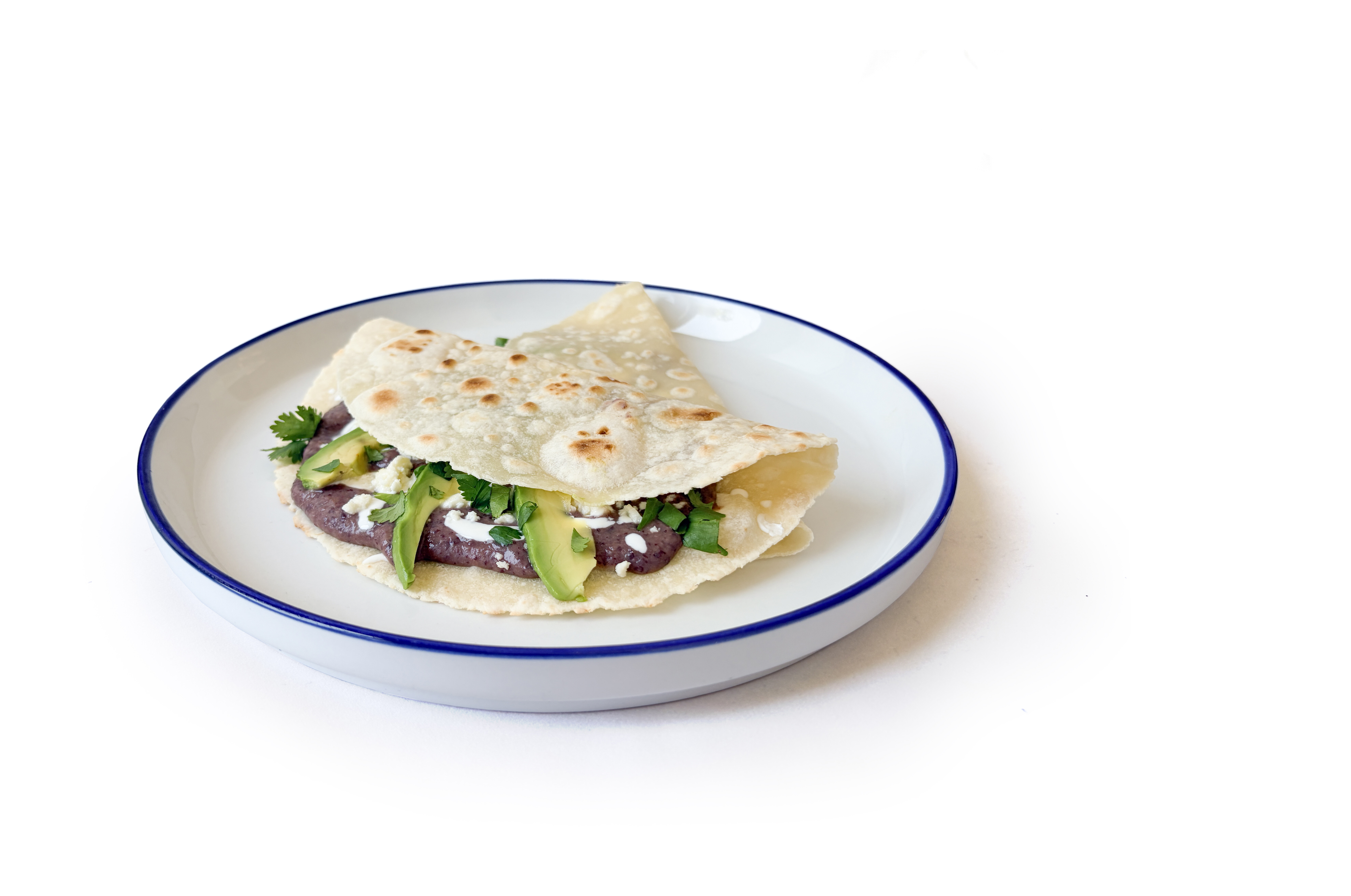 A tortilla folded on a white plate, filled with beans, avocado slices, cheese, and cilantro.