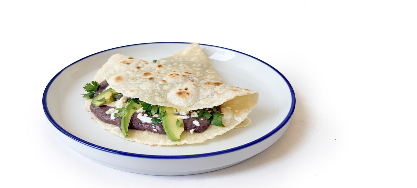 A tortilla folded on a white plate, filled with beans, avocado slices, cheese, and cilantro.