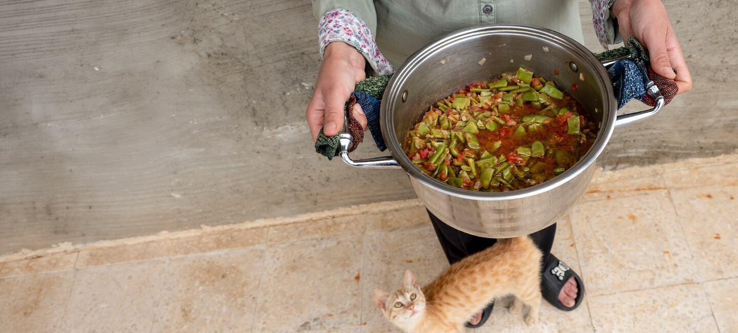 A person holds a pot of vegetable stew while a small orange kitten stands near their feet on a tiled floor.