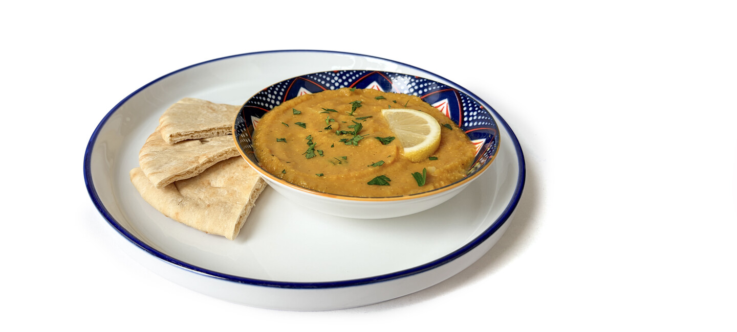 A bowl of lentil soup garnished with lemon and herbs is served with pita bread on a white plate against a white background.