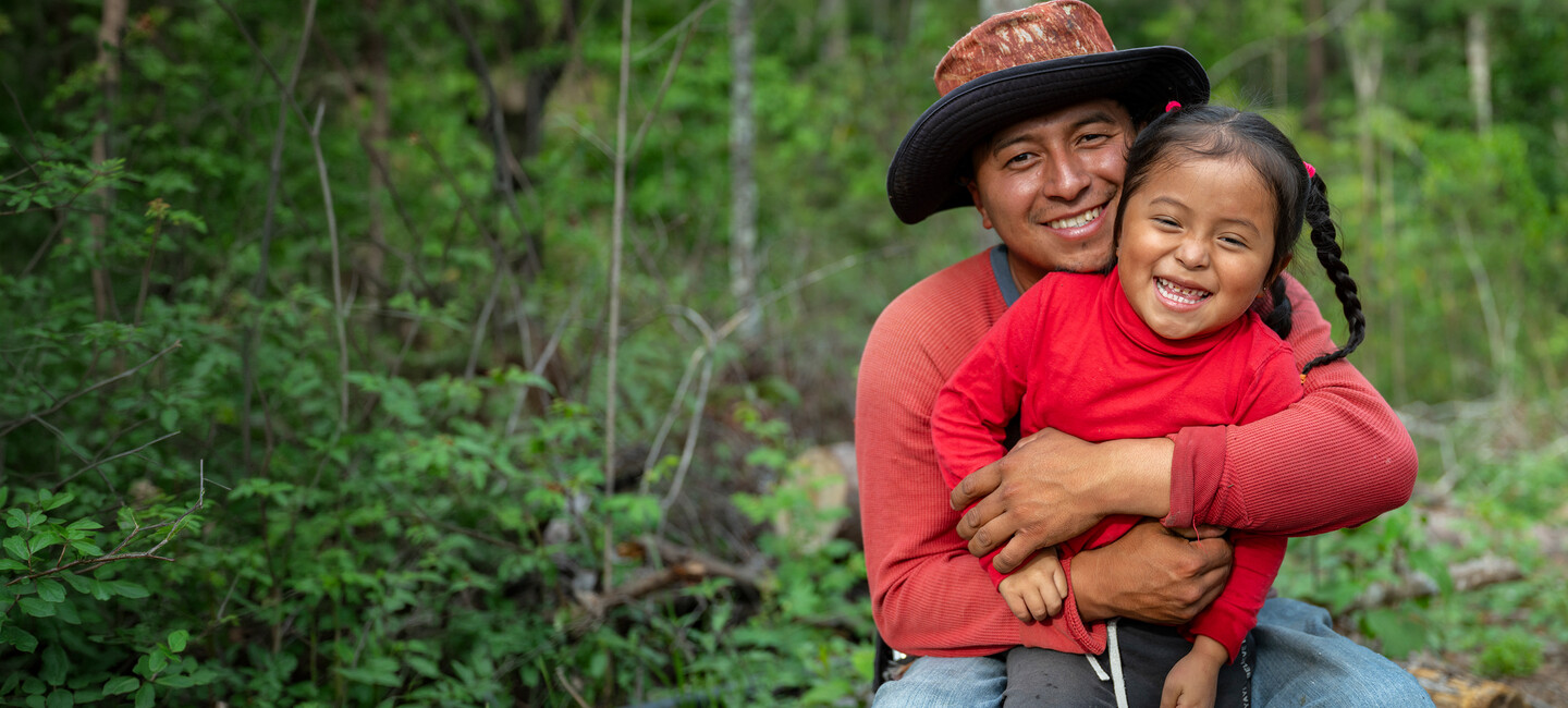 A man in a hat hugs a smiling young girl in red clothing outdoors, surrounded by green trees and vegetation.