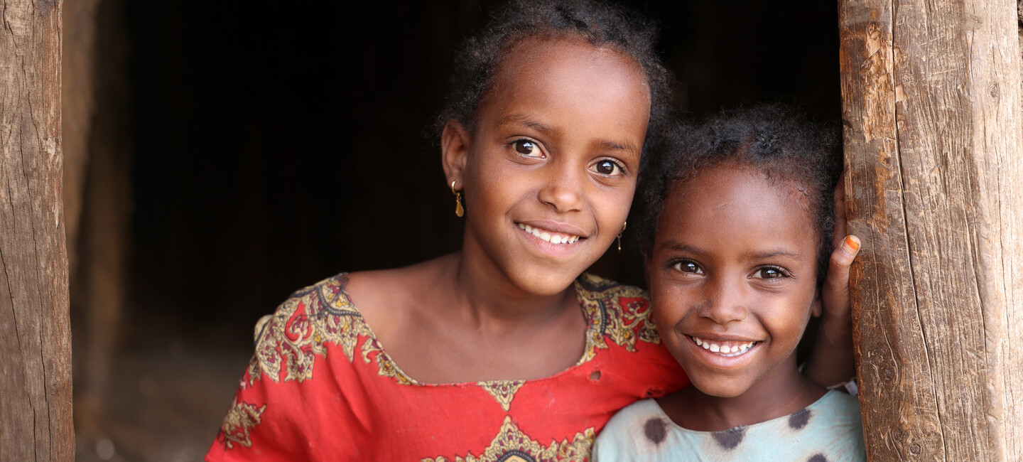 Two young girls stand close together in a doorway, smiling brightly at the camera.