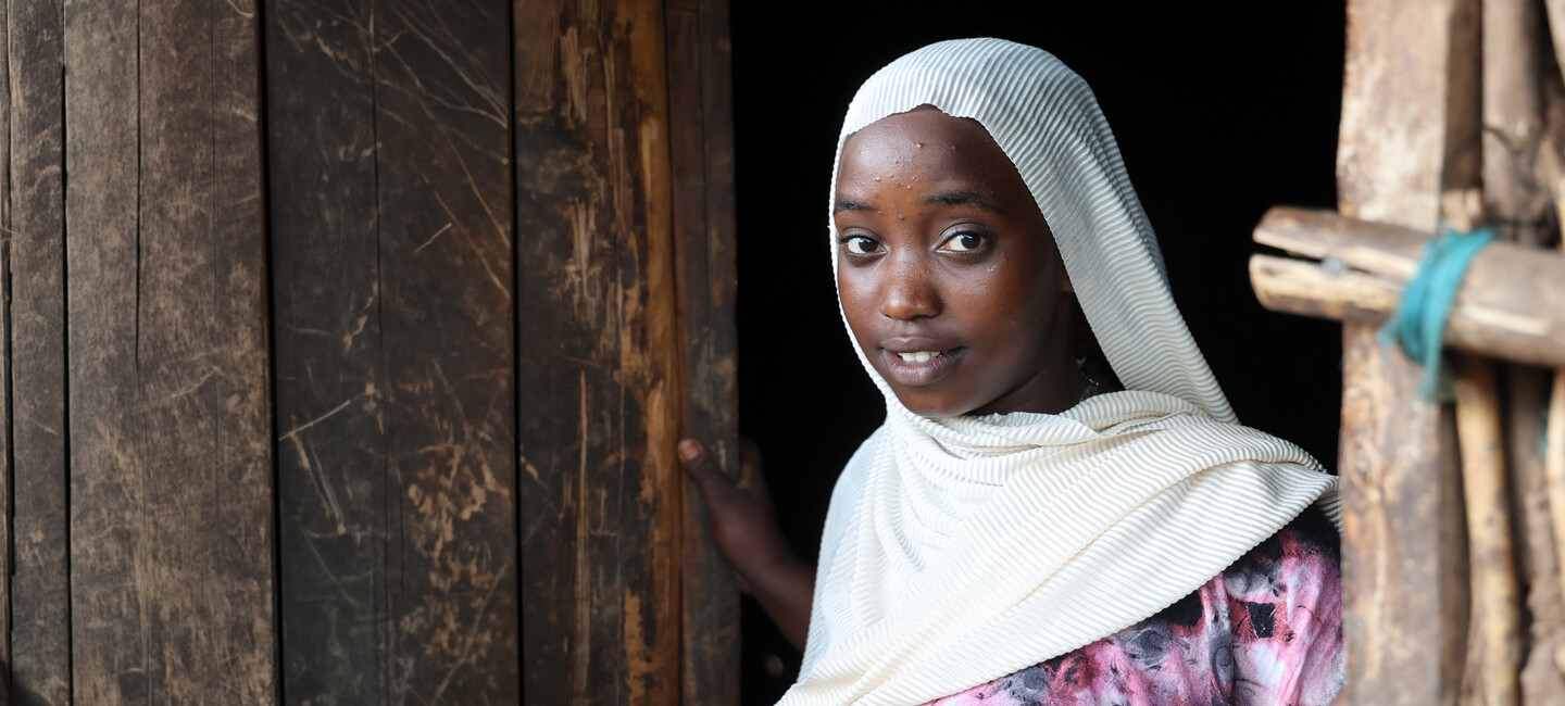 A girl wearing a white headscarf stands in a wooden doorway and looks toward the camera, smiling slightly.