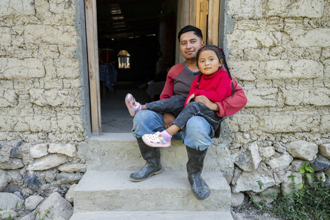 A man and a young girl sit together on the steps outside a rustic, mud-brick building, both looking at the camera.