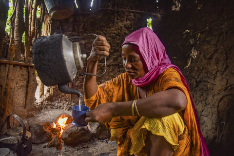 A woman in colorful clothing pours tea from a kettle into a cup inside a rustic kitchen with a fire in the background.