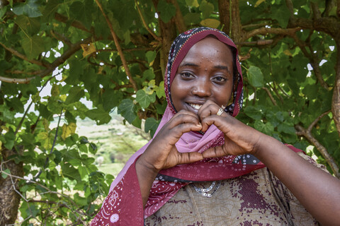 A woman wearing a patterned headscarf makes a heart shape with her hands while standing in front of leafy green trees.