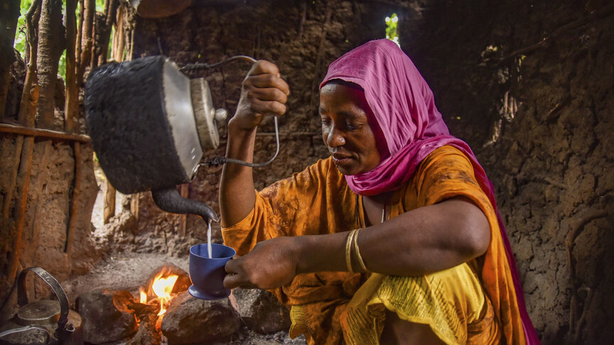 A woman in colorful clothing pours tea from a kettle into a cup inside a rustic kitchen with a fire in the background.